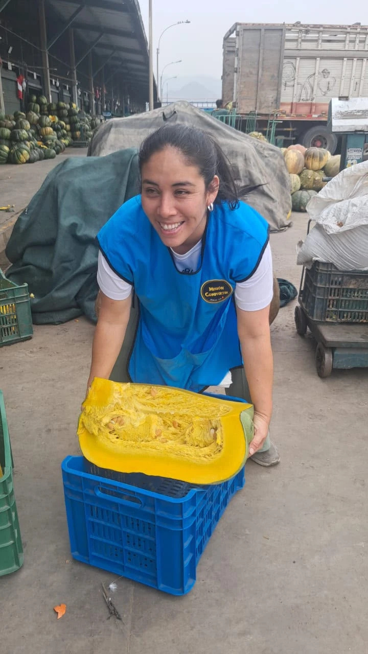 Una voluntaria de Misión Compartir con chaleco azul sonríe mientras carga medio zapallo sobre una caja azul en un mercado